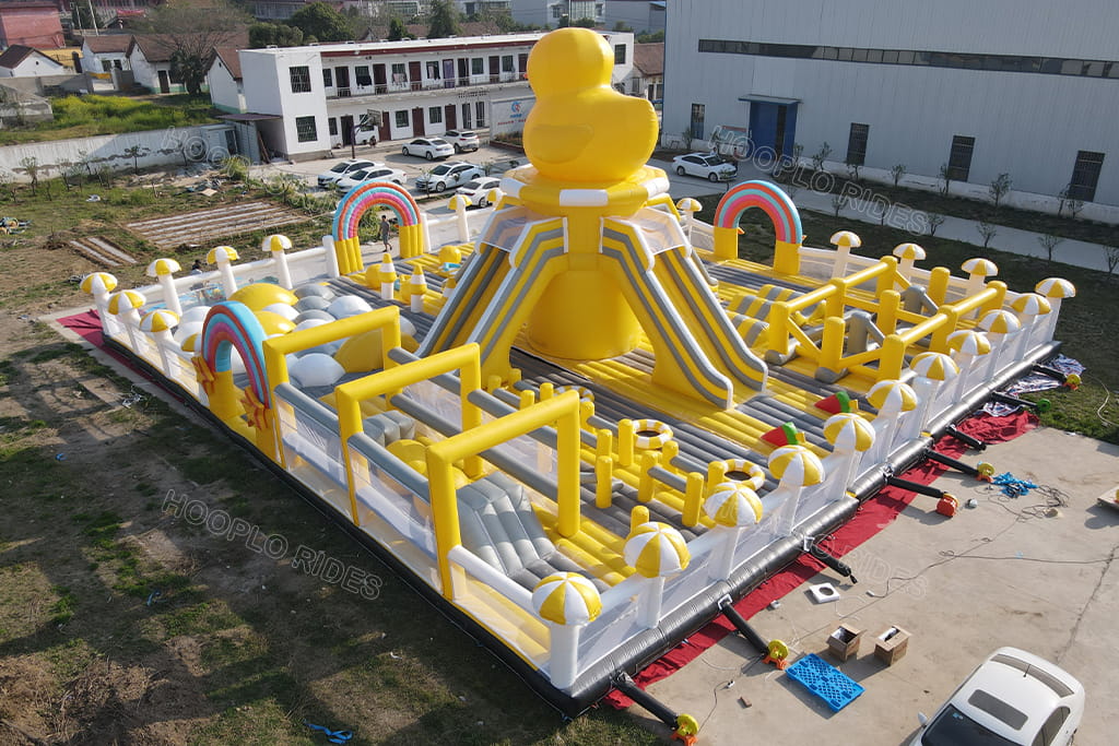 Side perspective of giant rubber duck inflatable obstacle course park, showing climbing challenge obstacles and mushroom‑shape fence decorations for outdoor kids event rental